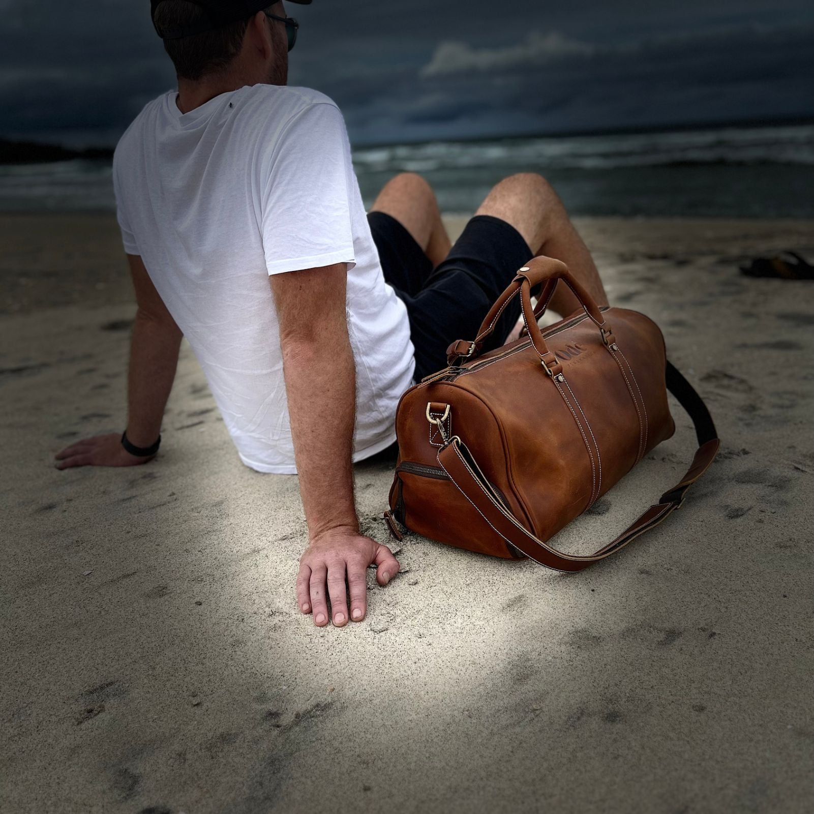 man in white t shirt sitting on the beach with his brown ode federation leather weekender bag